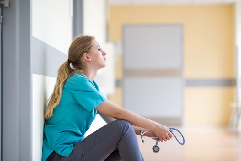 Tired and worried nurse sitting up against the wall holding stethoscope.